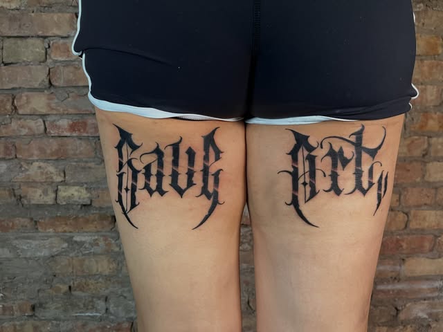 Bold black Gothic lettering tattoos across both thighs.