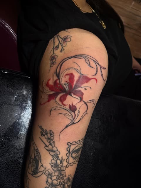 Red lily tattoo surrounded by fine-line flowers and leaves on an arm.