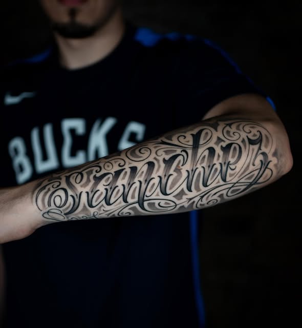 Ornate black script lettering tattoo across a forearm.